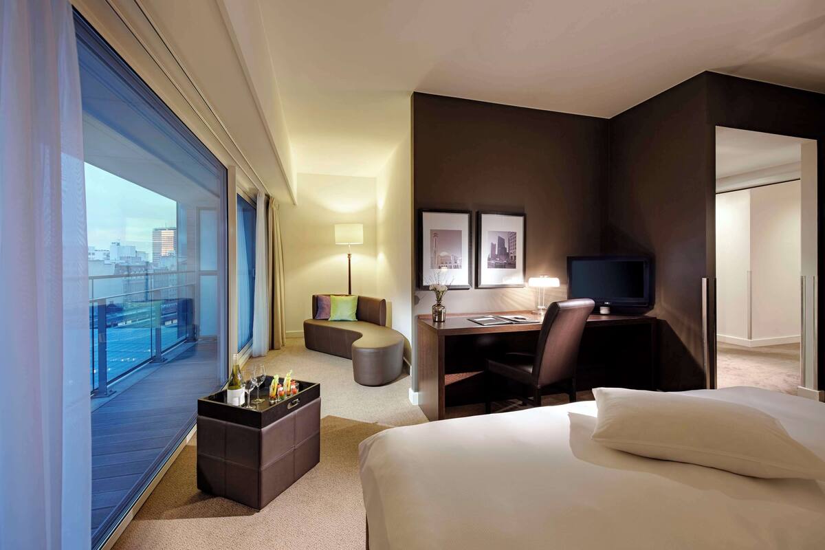 A hotel room with a large window, a bed, a desk with a chair, a sofa, and a television.