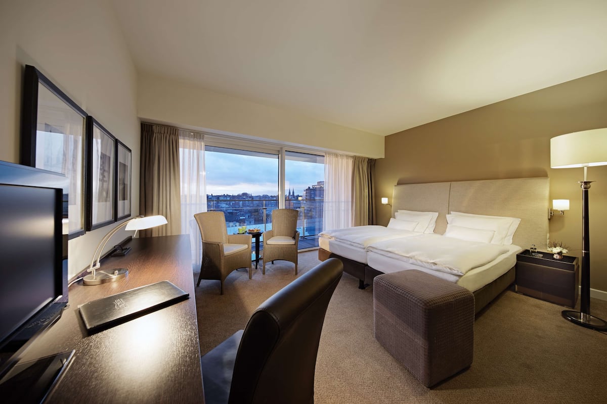 A hotel room with a bed, desk, chairs, a TV, and a view of the city.