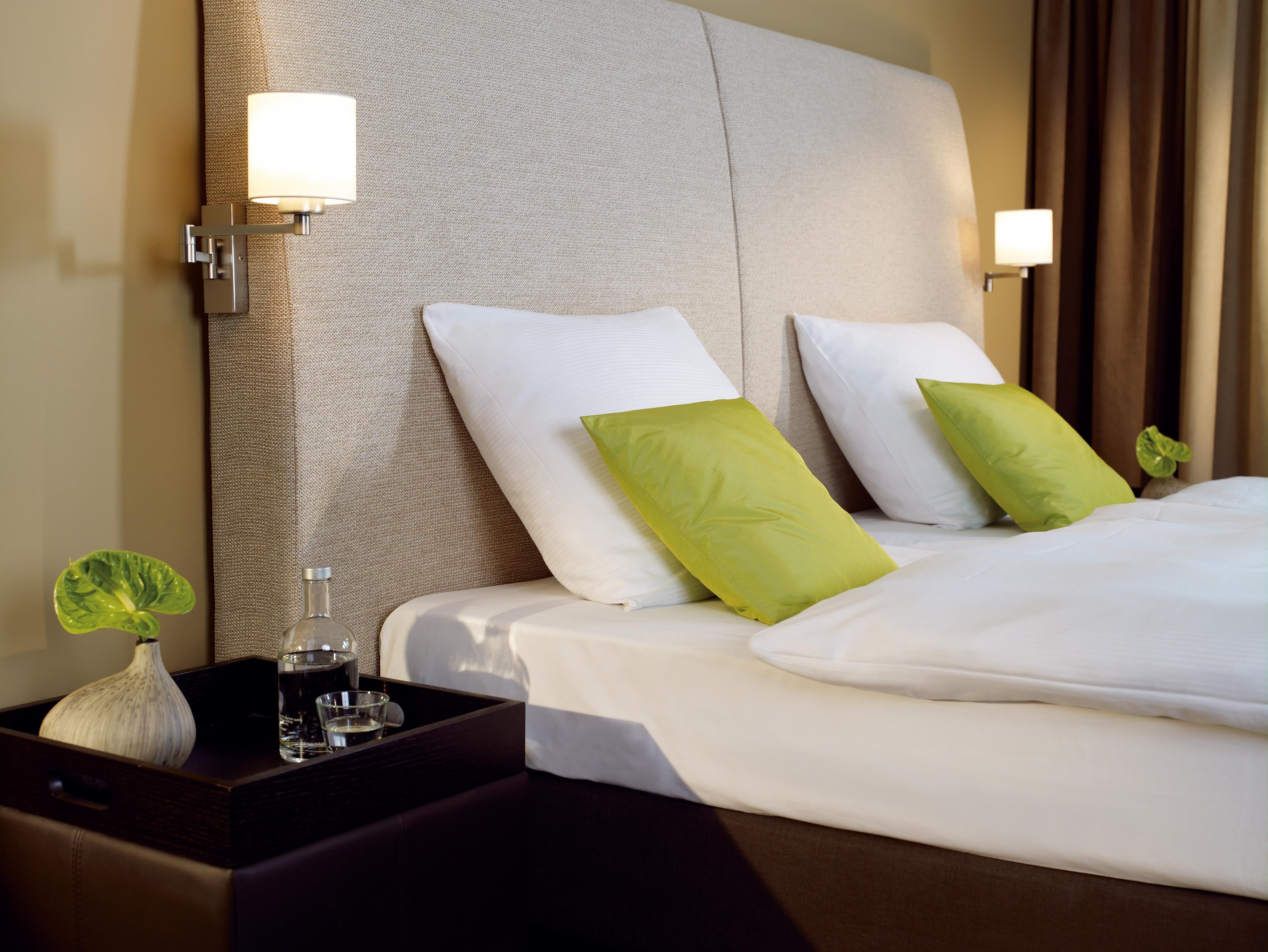A hotel room with a bed, pillows, bedside lamps, a nightstand with a vase and glasses, and a plant.