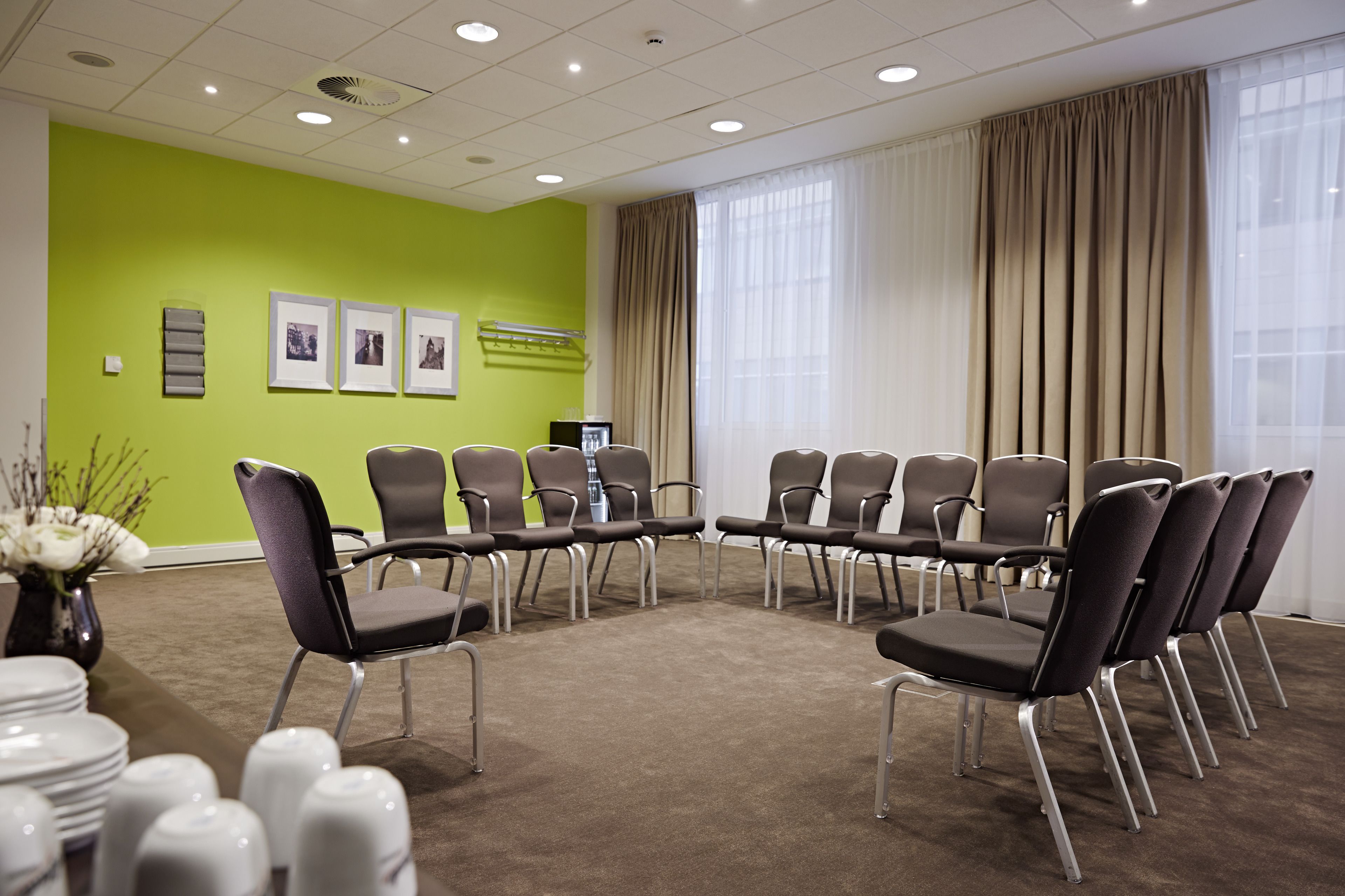 A conference room with a green wall, beige curtains, and a carpeted floor. There are several chairs arranged in a semi-circle, and a table with plates and glasses is visible.