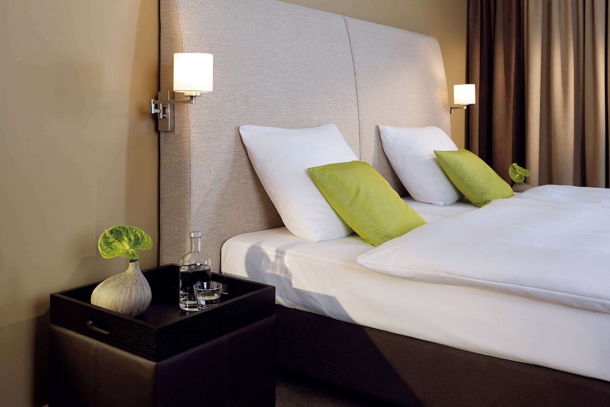 A hotel room with a large bed, two bedside tables, a nightstand, and a vase with a plant.