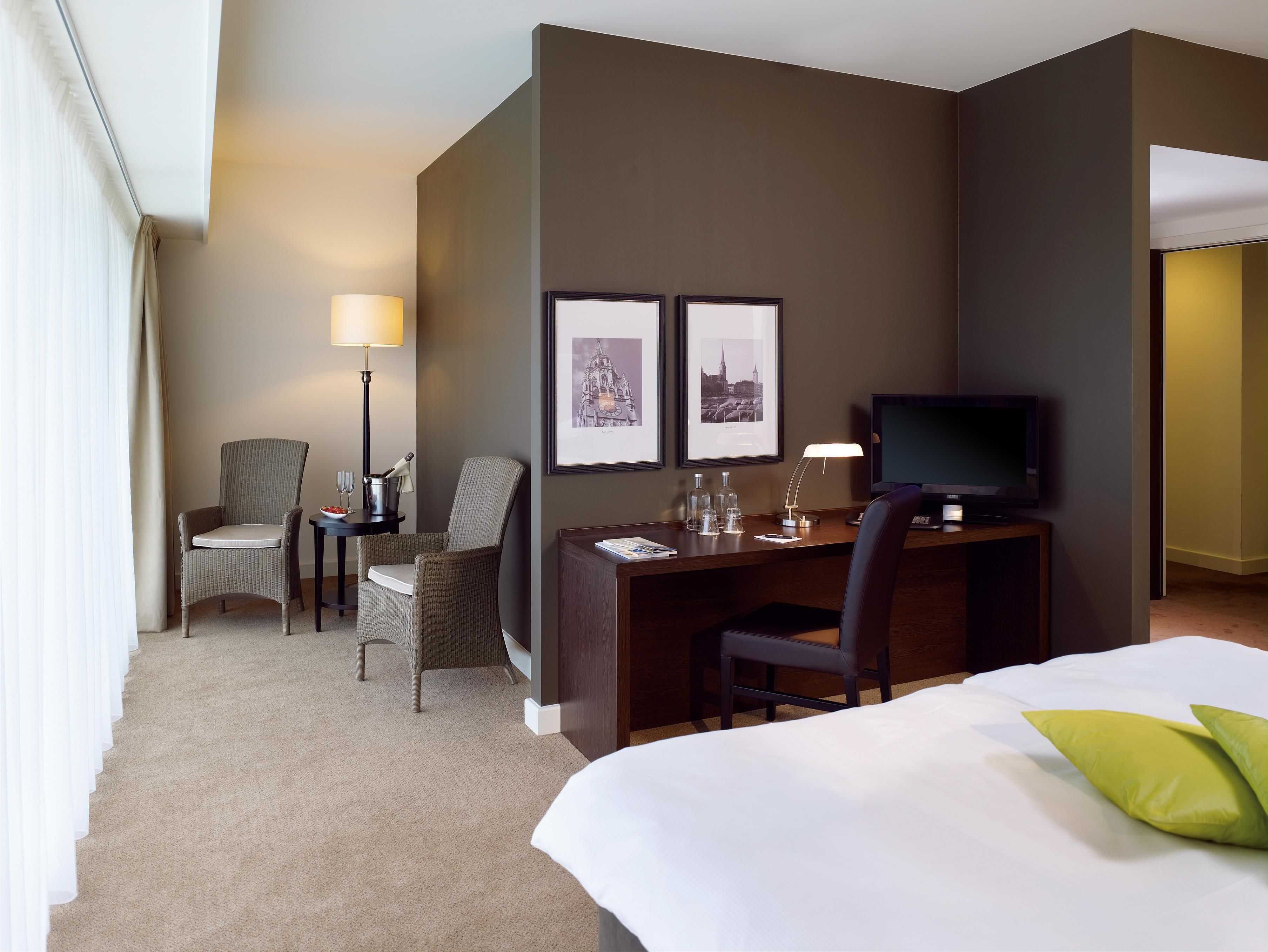 A hotel room with a bed, two chairs, a desk with a computer, a flat-screen TV, and framed pictures on the wall.