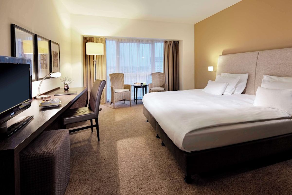 A hotel room with a large bed, a desk with a television, a chair, and a small table with a lamp.