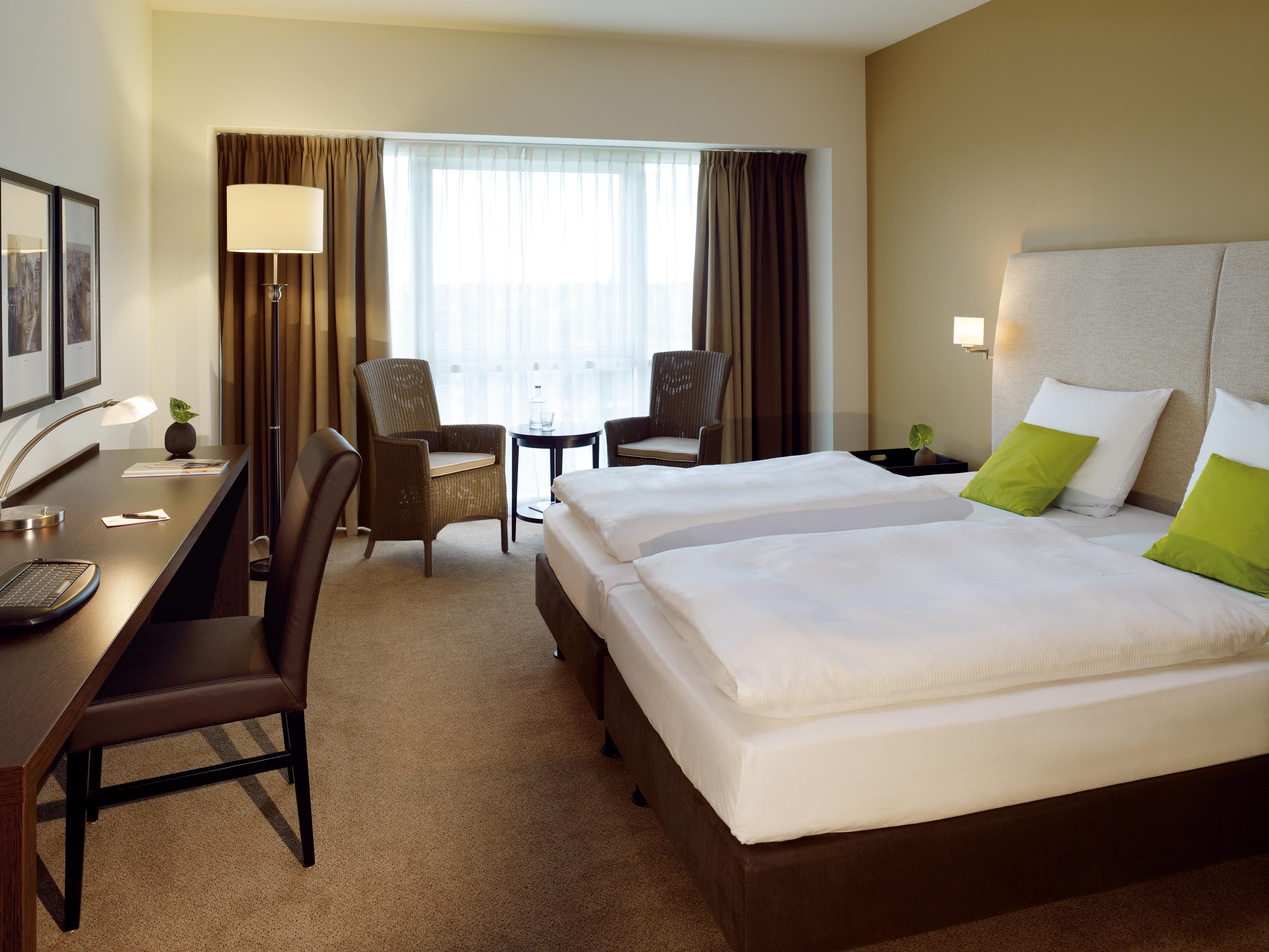 Ein Hotelzimmer mit Schreibtisch, zwei Stühlen, einem Bett mit weißer Bettwäsche und grünen Kissen, einer Lampe und einem Fenster mit Vorhängen.