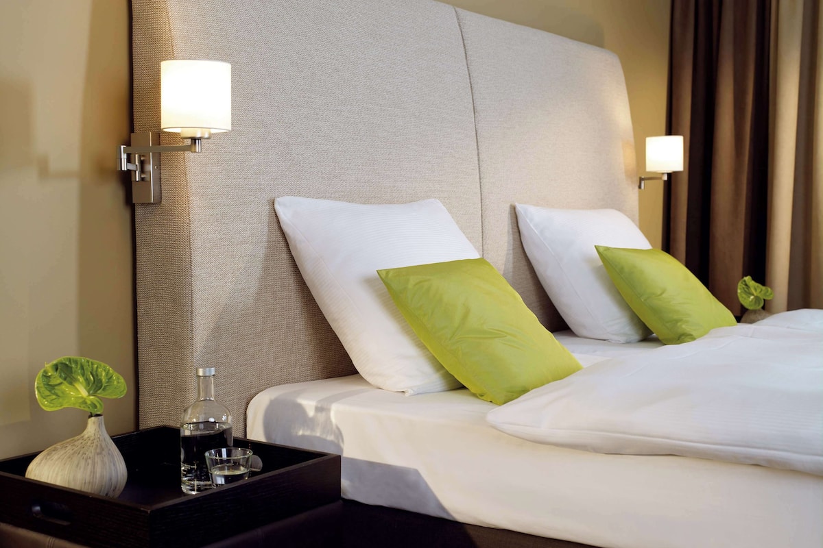 A neatly made bed with white and green pillows, a bedside table with a vase and glasses, and wall-mounted lamps.