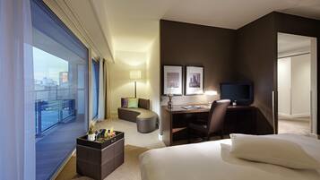 A hotel room with a bed, a desk, a chair, a TV, a sofa, and a balcony with a view of the city.