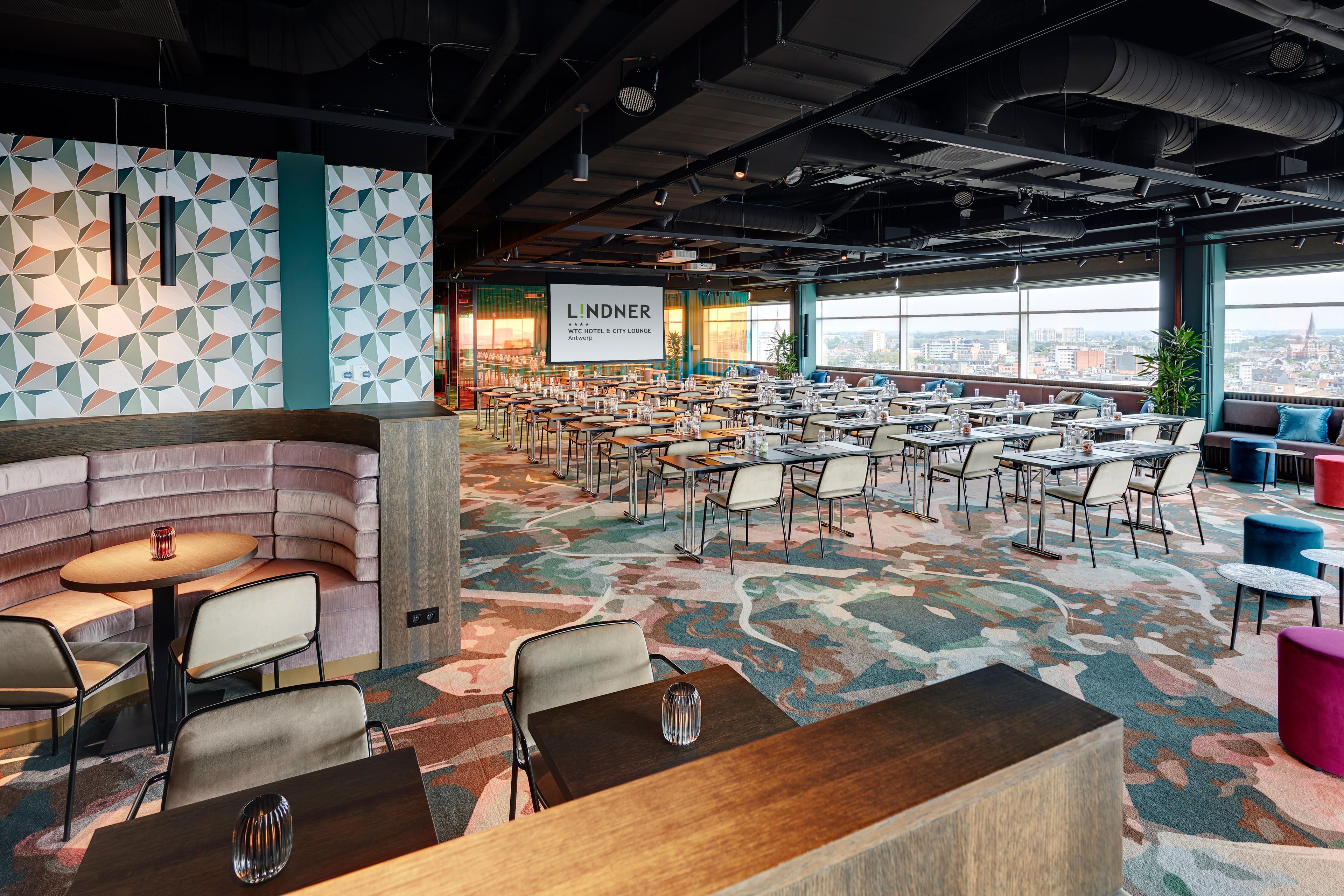 A spacious hotel lobby with a modern design, featuring a large seating area, multiple tables, and a city view through large windows.