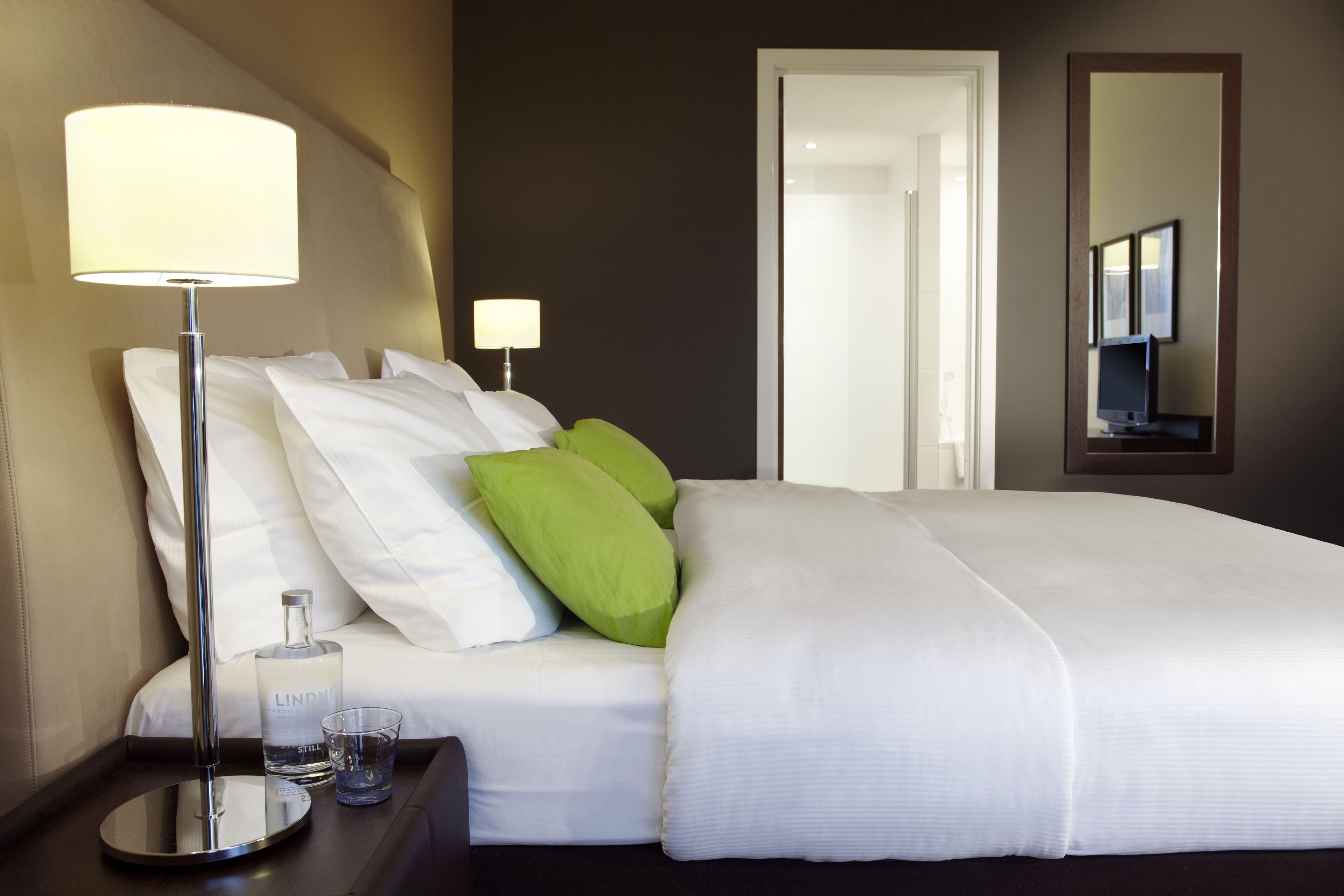 Quarto de hotel com cama, duas luminárias laterais, mesa de cabeceira com um copo de água e um espelho na parede.