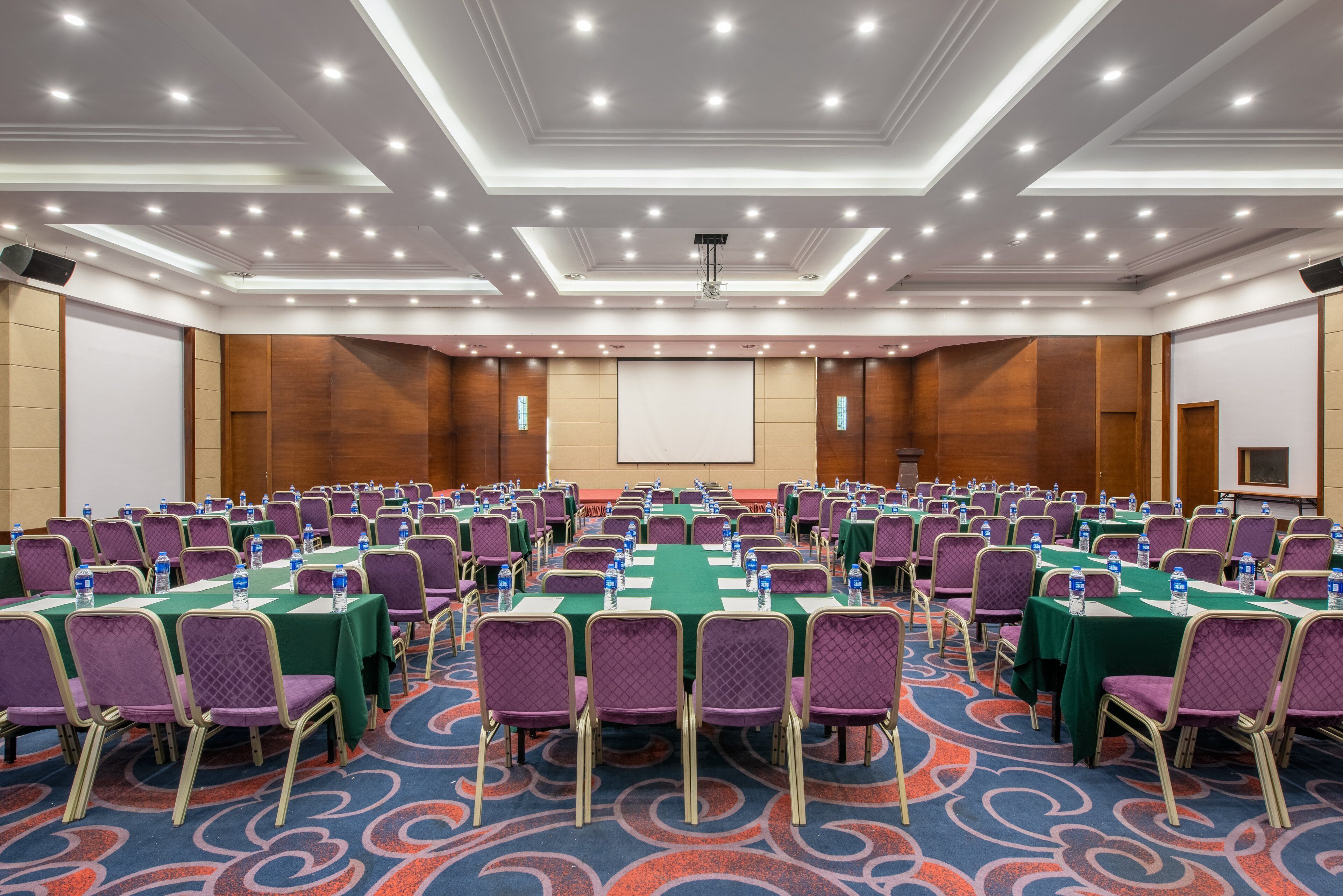 A conference room with rows of tables and chairs, equipped with water bottles and a projector screen.