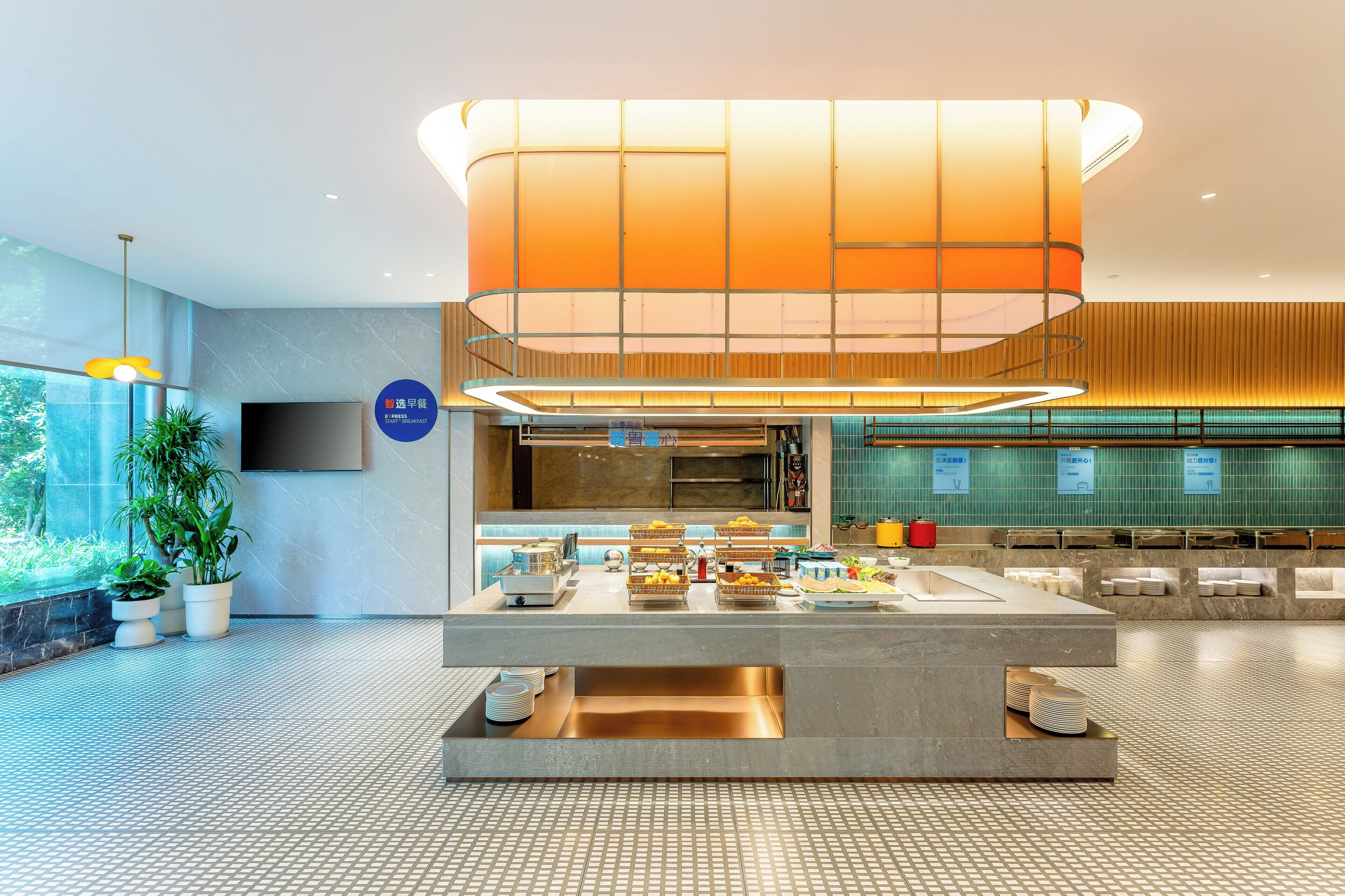 A modern hotel lobby with a central breakfast bar, a large hanging light fixture, and a television mounted on the wall.