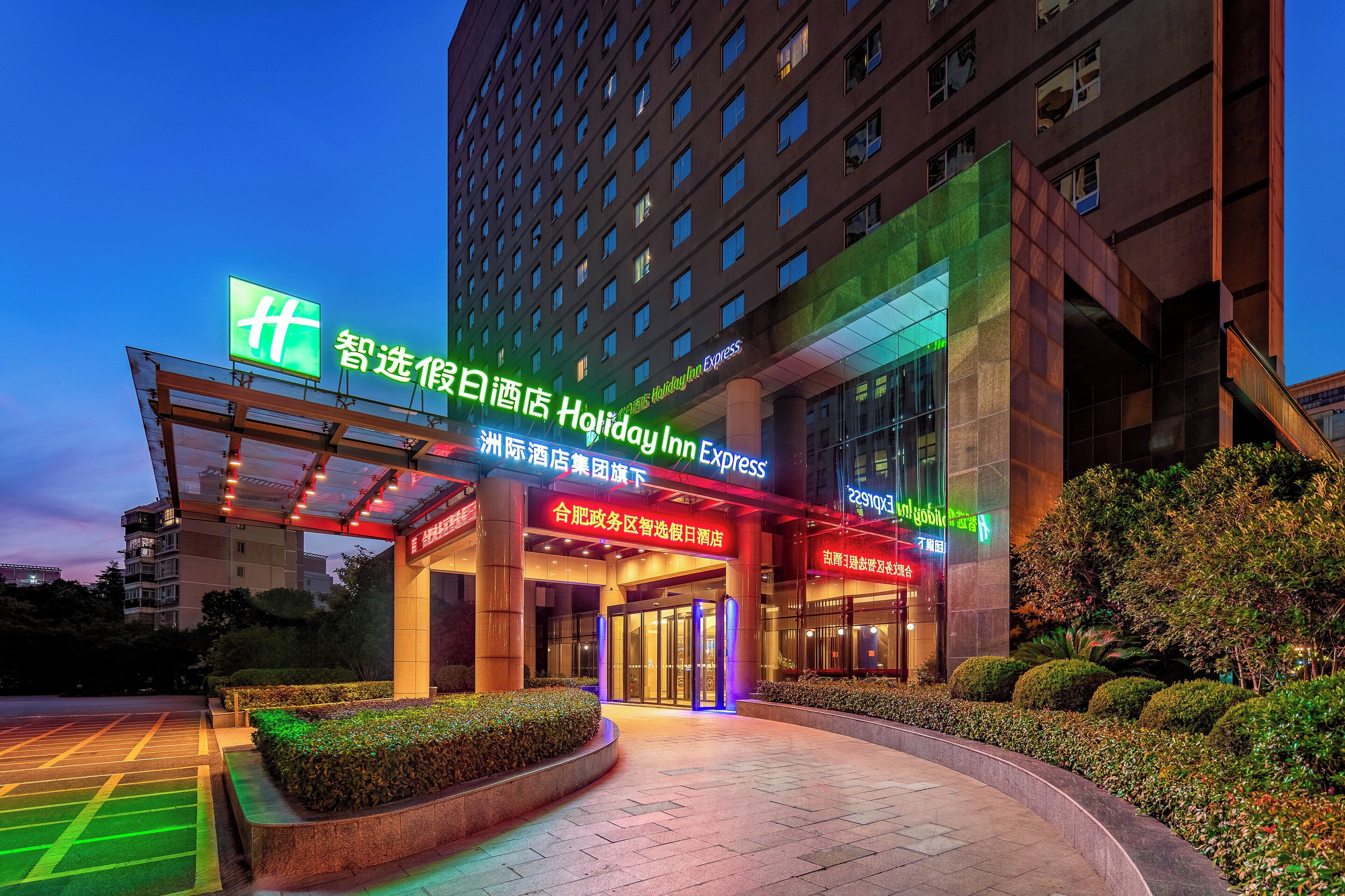 A Holiday Inn Express hotel entrance with a glass facade, illuminated signage, and a landscaped area with hedges.