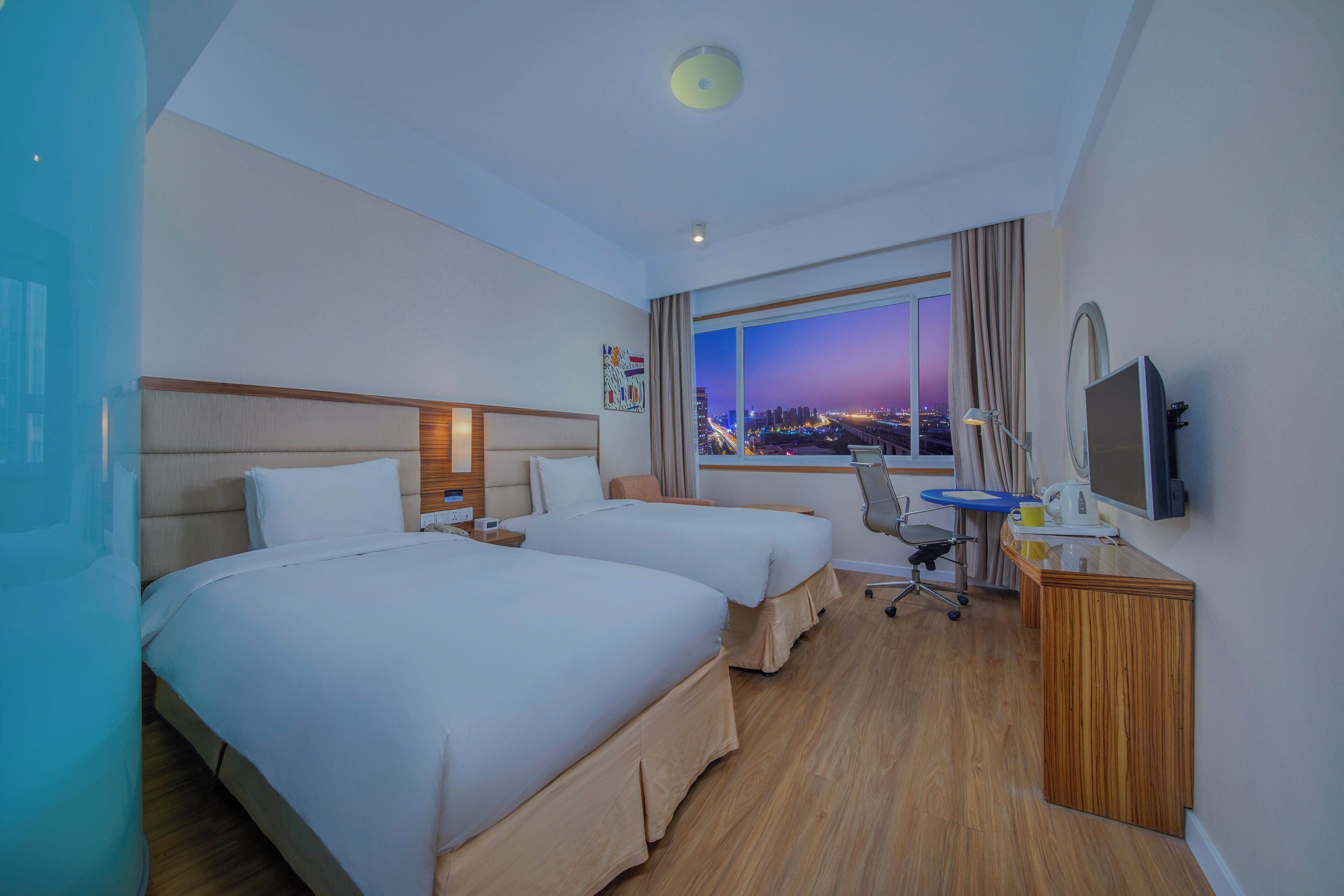 A hotel room with two beds, a desk, a chair, a TV, and a window with a city view.