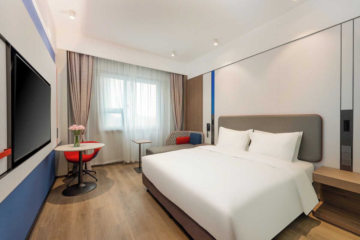 A modern hotel room with a large bed, a small table with a vase of flowers, a TV, and a window with curtains.