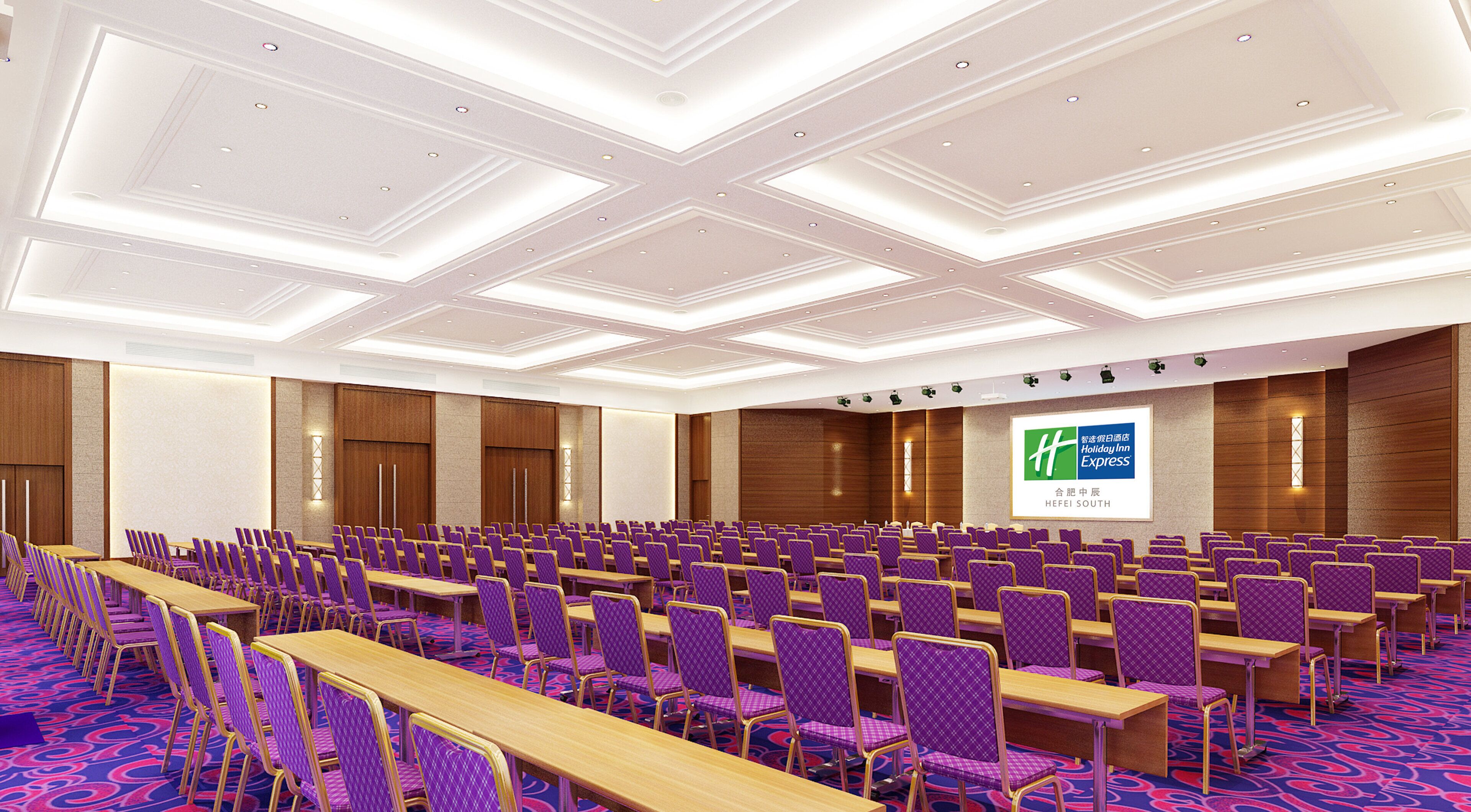 A large conference room with rows of purple chairs and wooden tables, a high ceiling with recessed lighting, and a Holiday Inn Express sign on the wall.