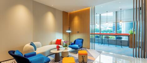 A modern hotel lobby with a sofa, chairs, ottomans, a coffee table, and a view of the city through large windows.