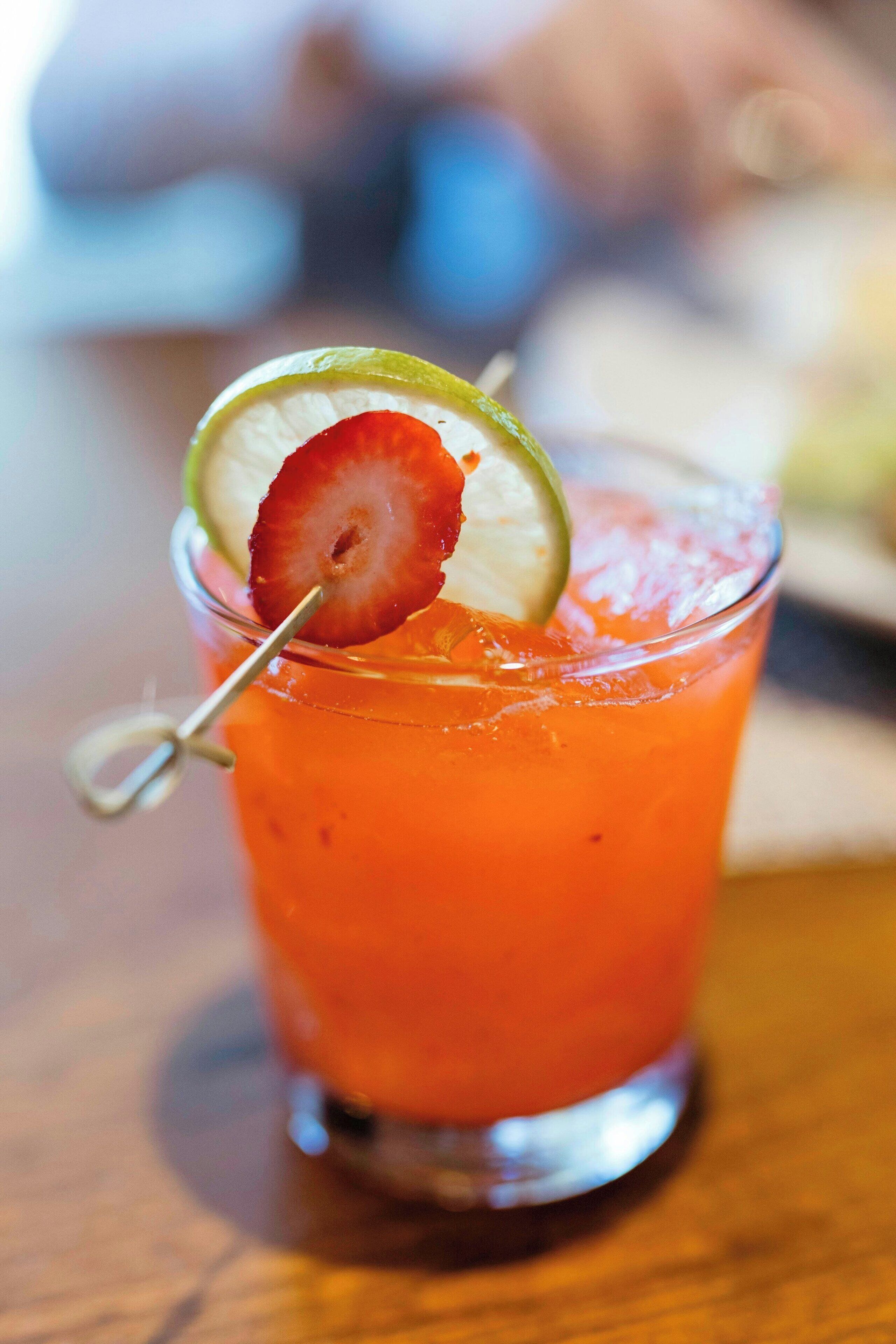 A glass of red beverage with a strawberry slice and a lime wedge on a wooden table.