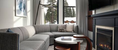 Un salon moderne avec un canapé d’angle, une cheminée et une grande fenêtre donnant sur des arbres et un chalet.