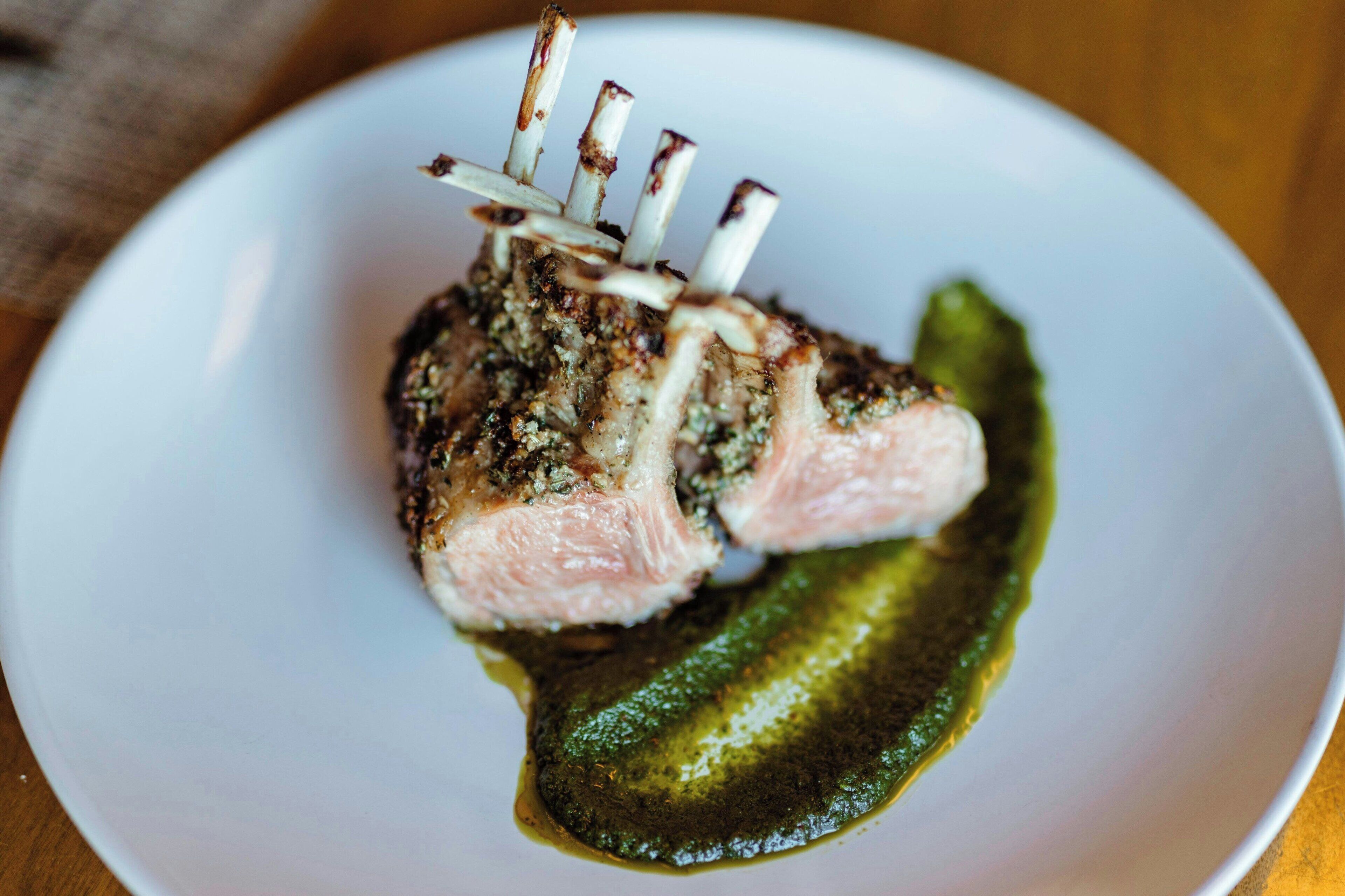 Une assiette contenant un morceau de viande, peut-être de l’agneau, recouvert d’une croûte aux herbes et accompagné d’une sauce verte.