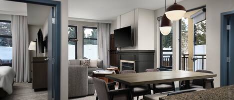 A modern hotel room with a dining area, a flat-screen TV mounted above the fireplace, and a large window offering a view of the outdoors.