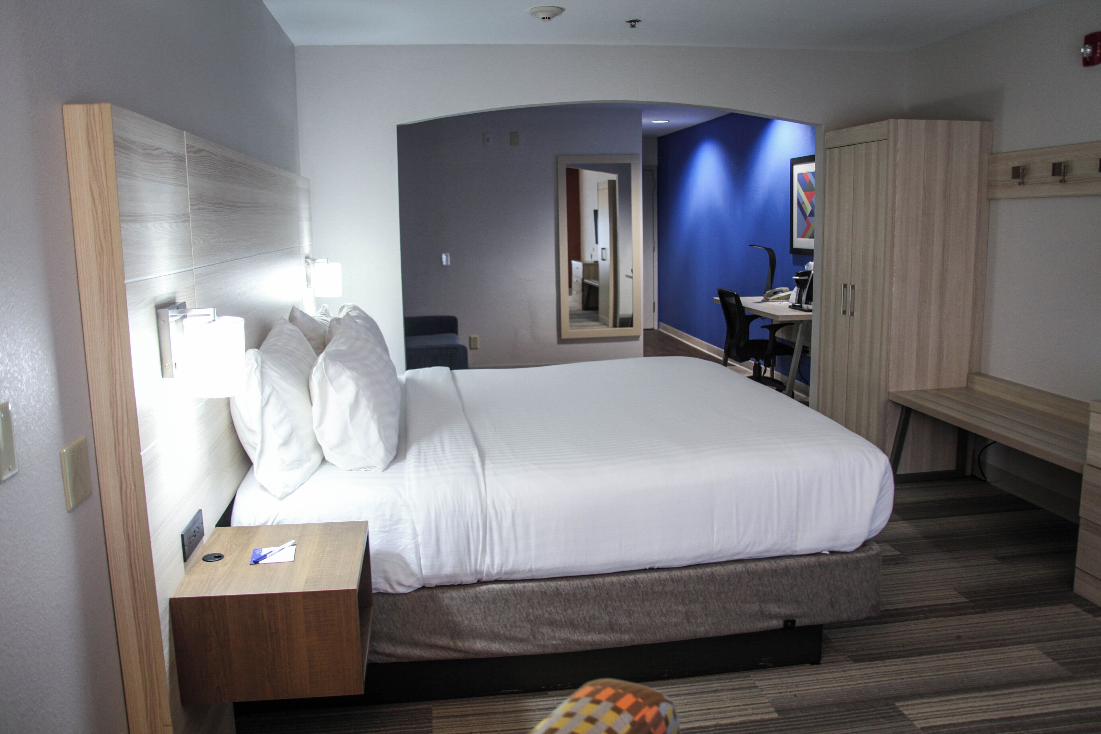 A hotel room with a large bed, a desk with a lamp, a wardrobe, and a bathroom visible through an archway.