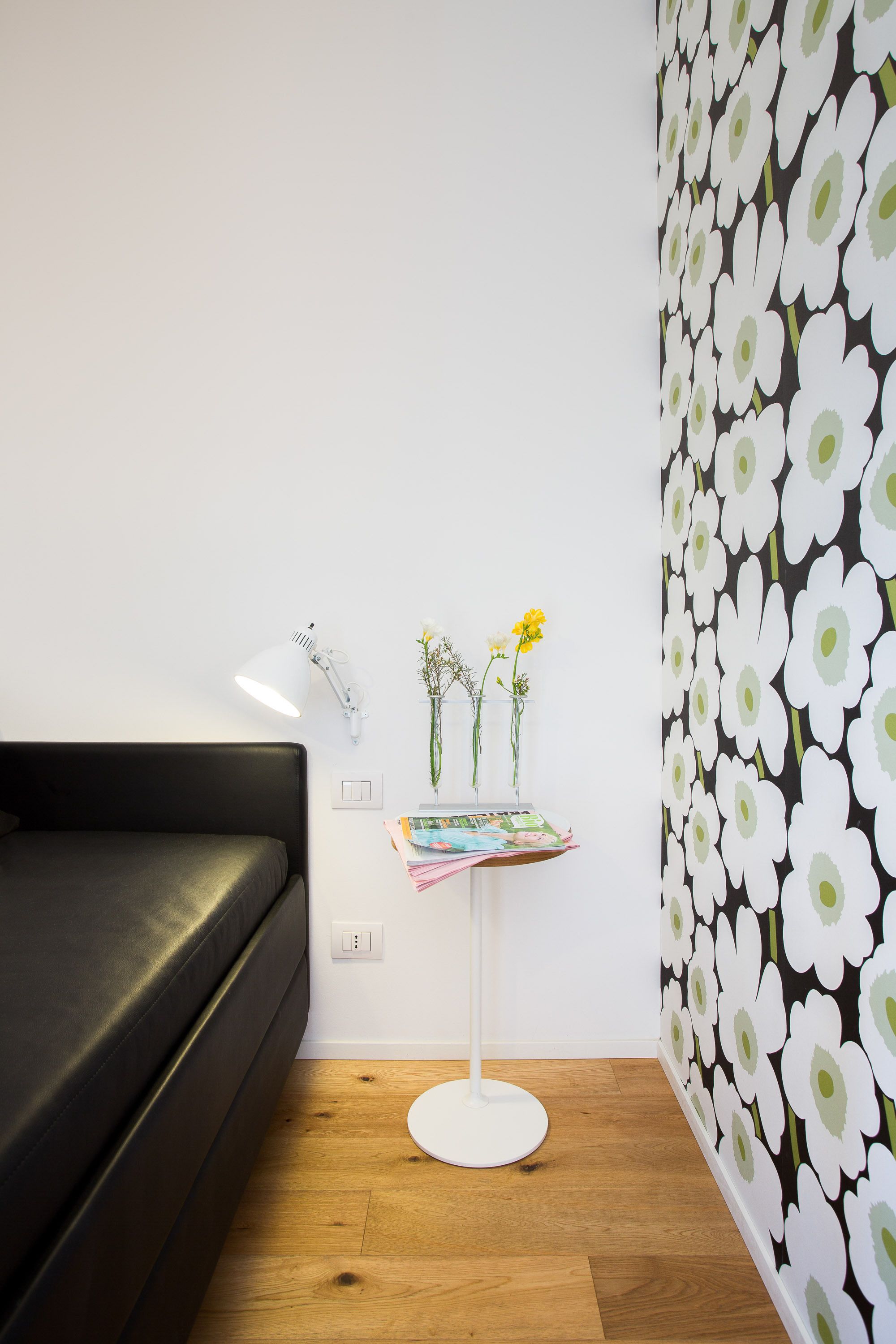 Una stanza moderna con un divano nero, una lampada bianca, un tavolino con dei fiori e una parete decorata con un motivo floreale.