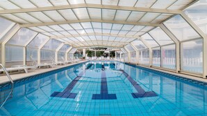 Una amplia piscina interior con techo de cristal, rodeada de sillones de descanso y vistas a edificios exteriores.