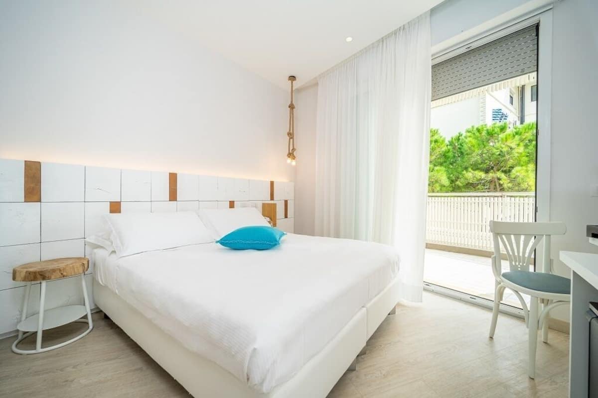 Un dormitorio moderno con una cama grande, un taburete de madera, una silla blanca y vistas al exterior con vegetación.
