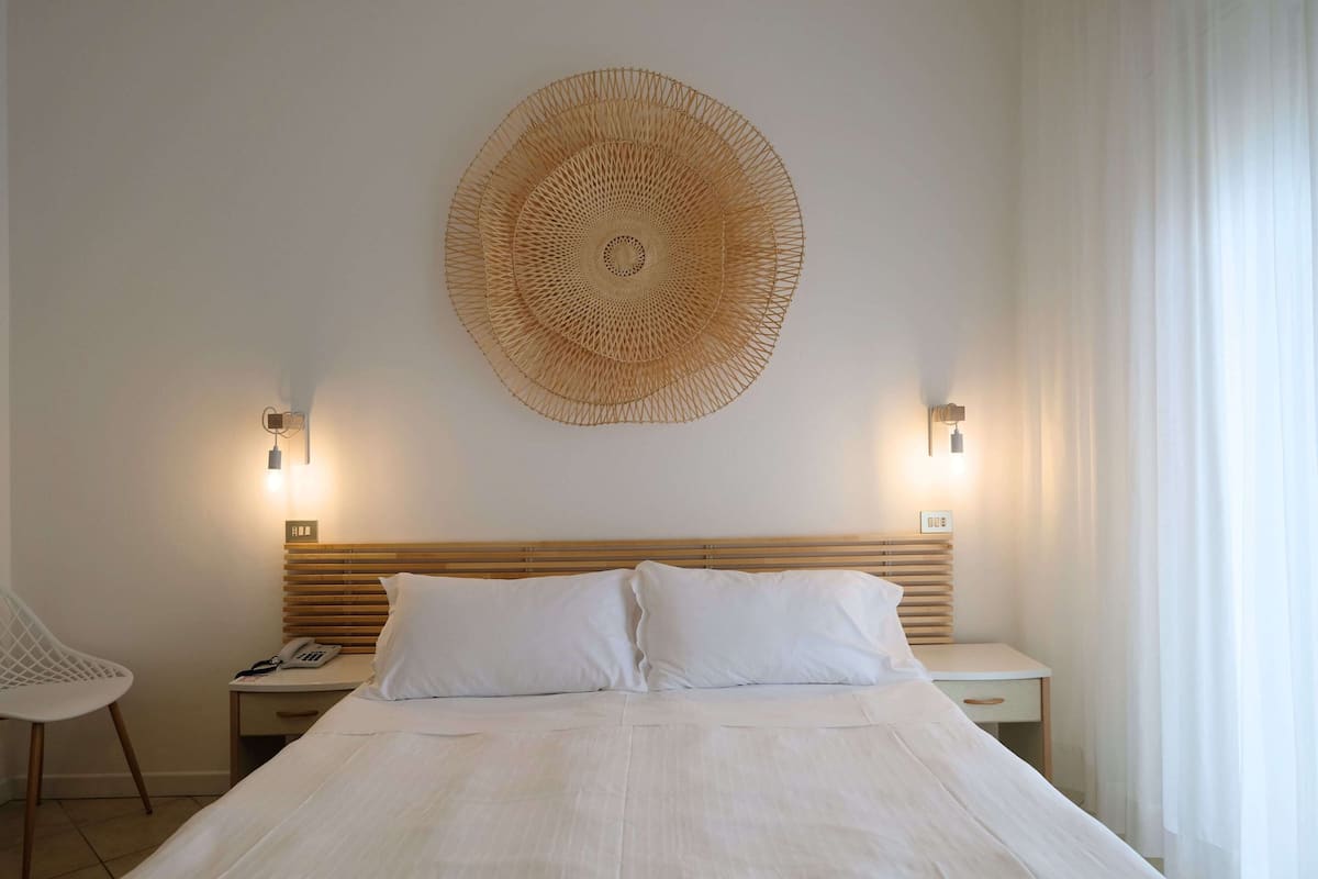 Una habitación de hotel ordenada con una cama grande, dos mesitas de noche, una silla y un adorno en la pared.