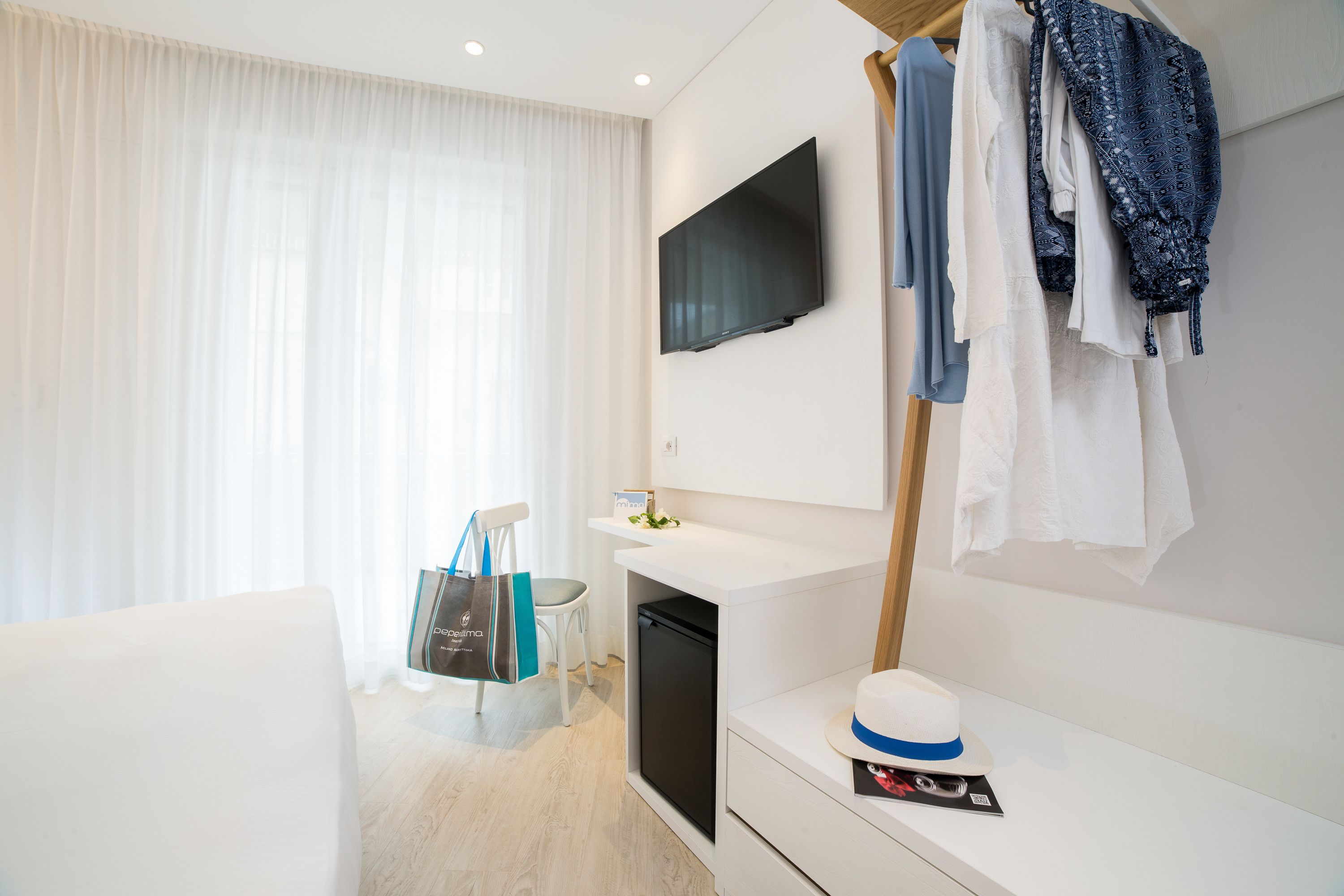 A modern hotel room with a flat-screen TV mounted on the wall, a small refrigerator, a white desk with a hat, and a clothing rack with various garments.