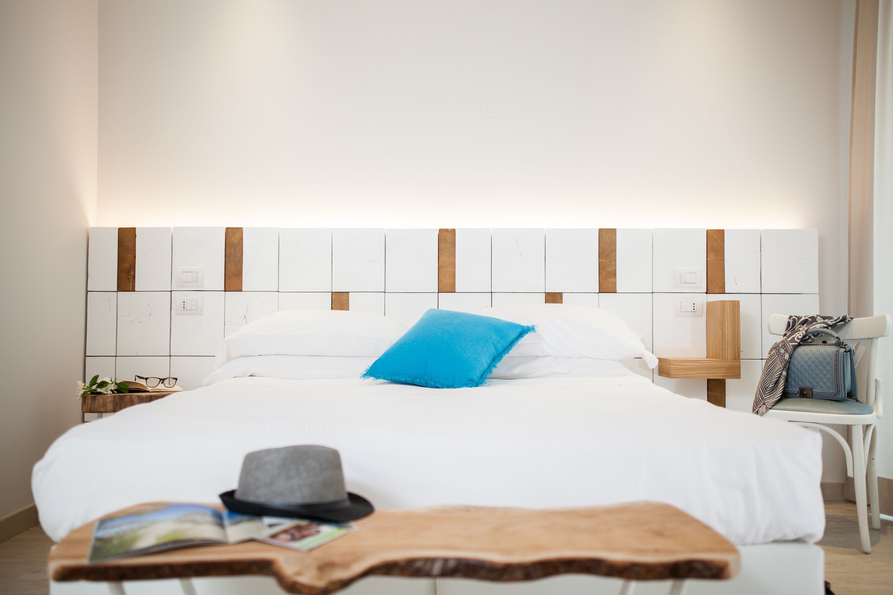 A neatly made bed with white bedding and a blue pillow, a wooden bedside table with a magazine and a hat, and a chair with a patterned bag.