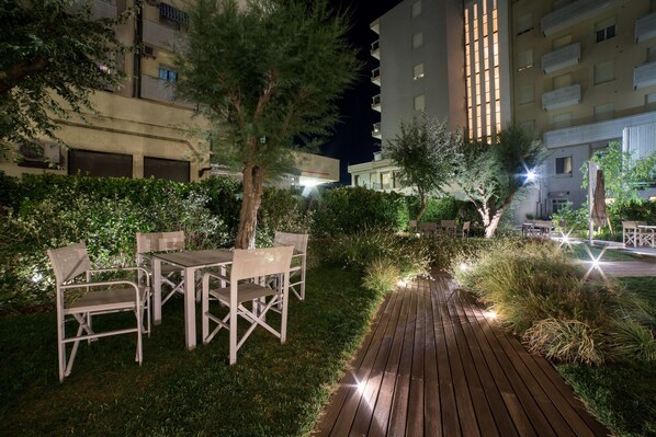 An outdoor patio area with wooden tables and chairs, surrounded by greenery and a wooden pathway.