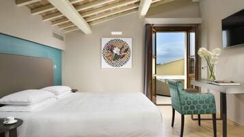 A hotel room with a bed, a desk with a chair, a painting on the wall, and a view through an open door.