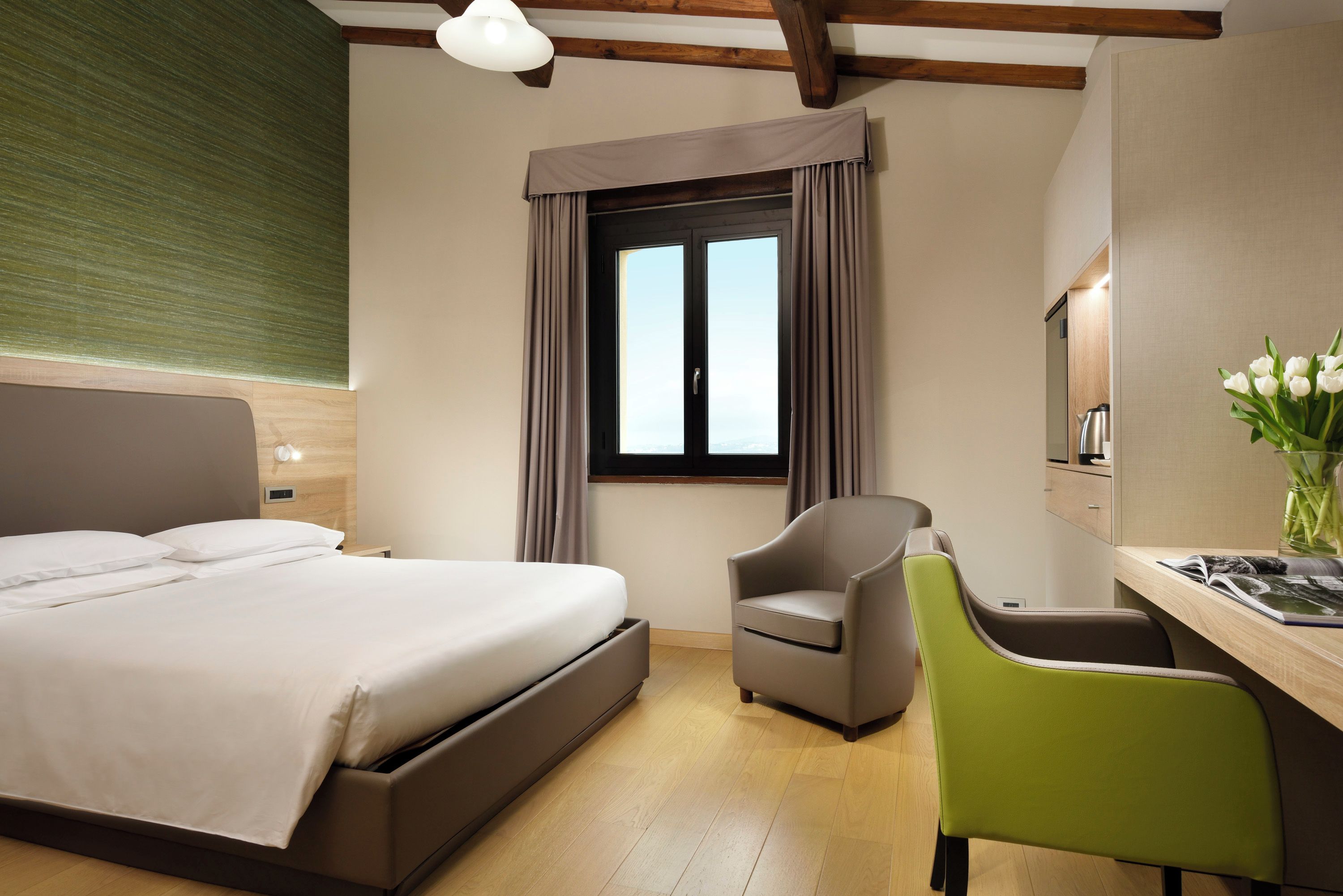 A hotel room with a bed, a desk with a chair, a window with curtains, and a vase with flowers.