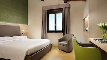 A hotel room with a bed, a desk with a chair, a window with curtains, and a vase with flowers.