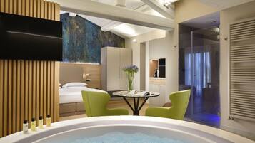 A modern hotel room with a large bathtub, a flat-screen TV, a wooden panel wall, a bed, a small table with a vase, and a shower area.