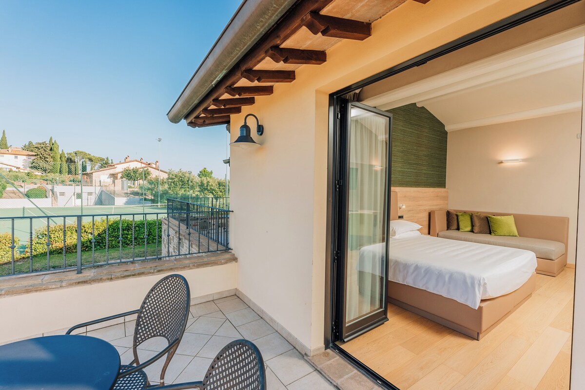 A balcony with a table and chairs, a bed with a headboard, and a view of houses in the distance.