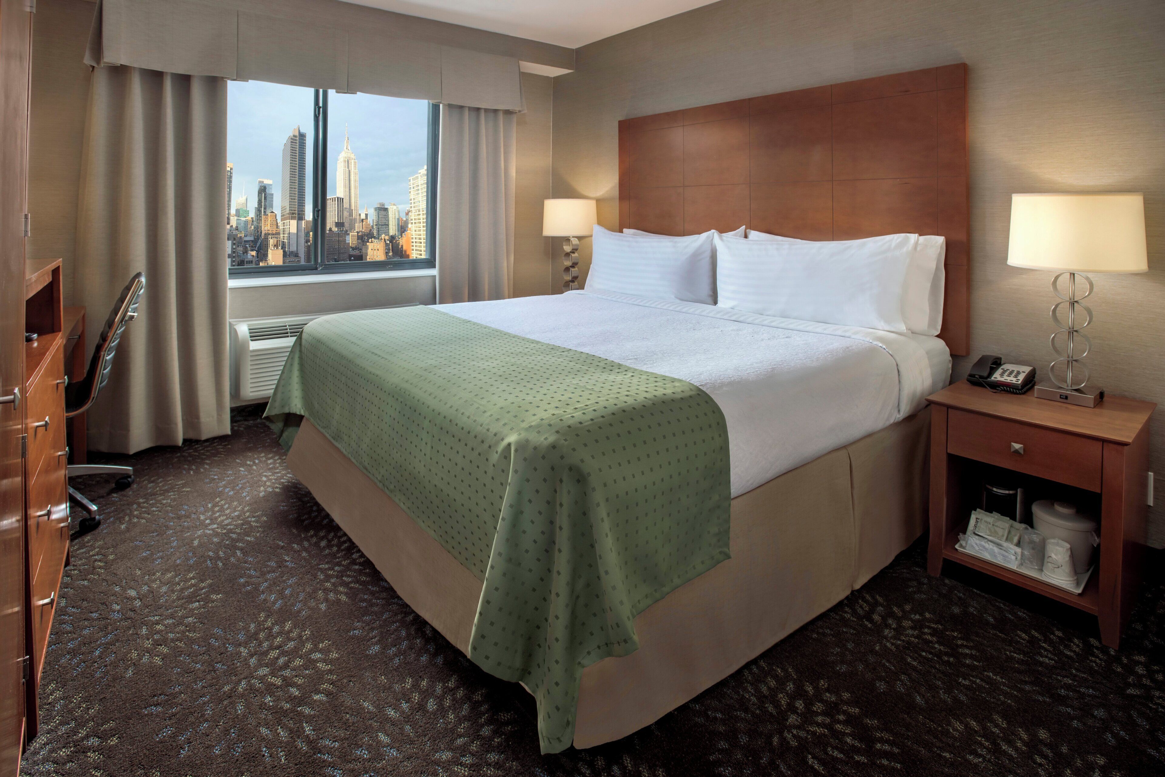 A hotel room with a bed, a desk, a chair, a nightstand with a lamp, and a view of the city skyline.