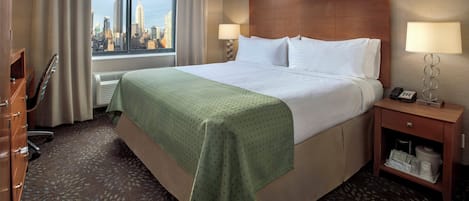 A hotel room with a bed, a desk, a chair, a nightstand with a lamp, and a view of the city skyline.