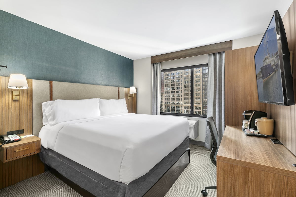 A hotel room with a large bed, bedside tables, a desk with a coffee maker, a TV, and a window with a city view.