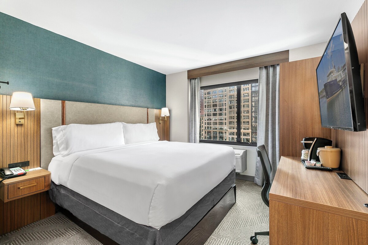 Une chambre d’hôtel avec un grand lit, des tables de chevet, un bureau avec une machine à café, une télévision et une fenêtre donnant sur la ville.