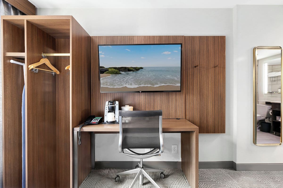 A compact hotel room with a wooden desk, a chair, a television mounted on the wall, a mirror, and a wardrobe with a hanging rack.