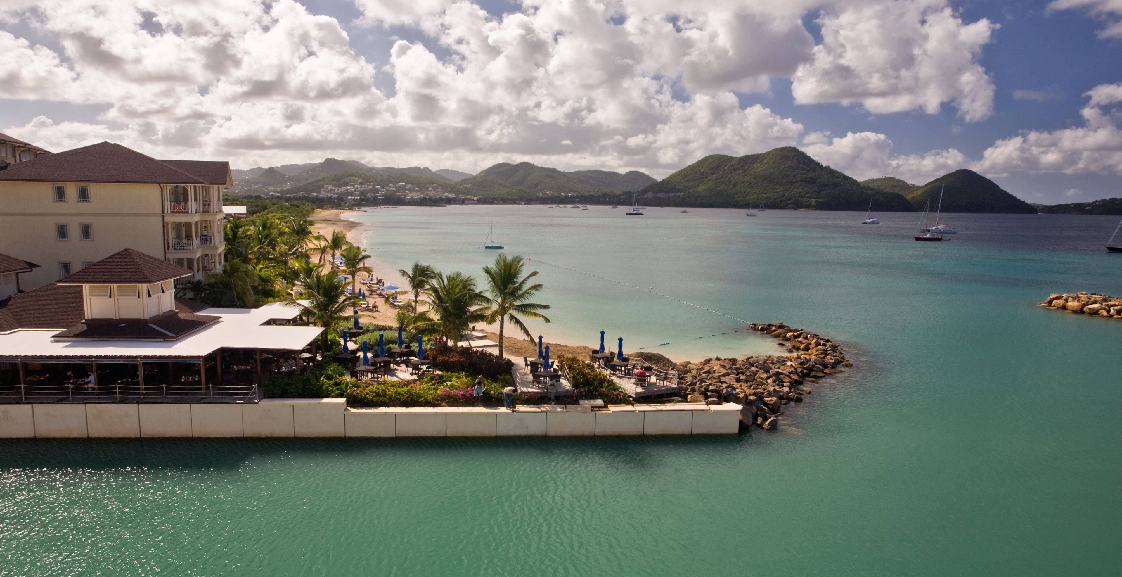 Un hotel frente al mar con playa, palmeras y barcos al fondo.