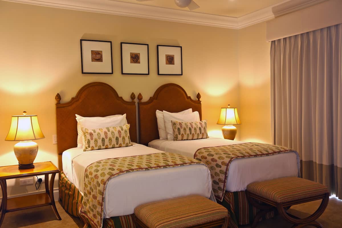 A hotel room with two beds, a nightstand with a lamp, a window with curtains, and framed pictures on the wall.