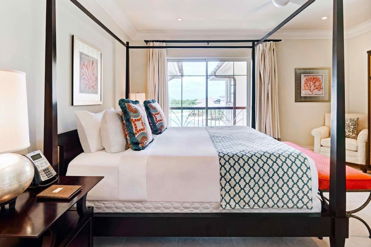 A four-poster bed with a view of the outdoors, a bedside table with a lamp, and a chair in the corner.