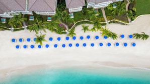An aerial view of a beach with rows of blue umbrellas and white lounge chairs, surrounded by palm trees and a swimming pool. - The Landings Resort & Spa (Gros Islet)