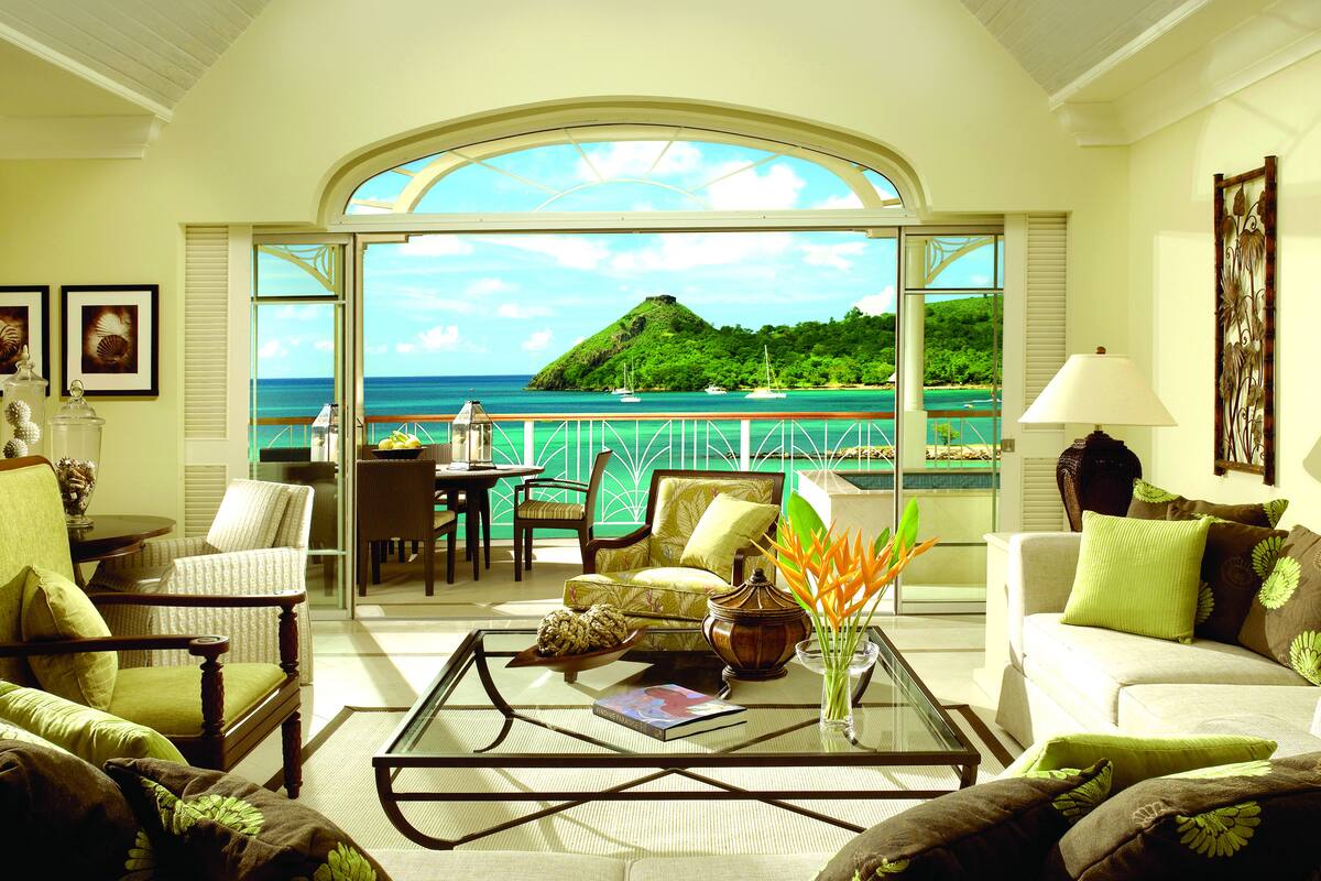 A spacious living room with a large glass door leading to a balcony with a view of the sea and a hill.