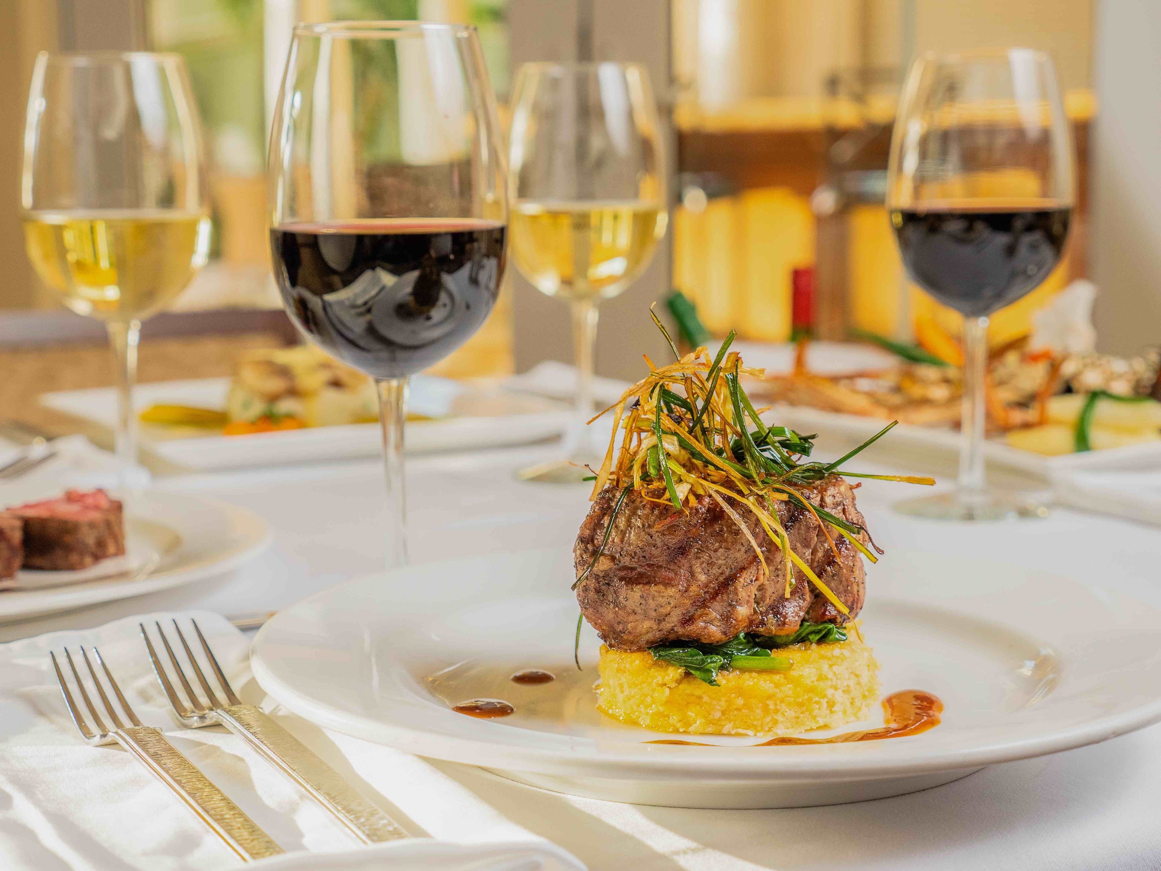 A plated dish with a steak, garnished with green herbs and thinly sliced vegetables, accompanied by a glass of white wine and a glass of red wine.
