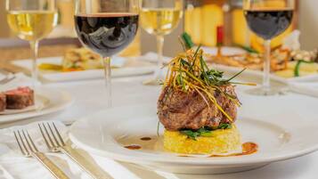 A plated dish with a steak, garnished with green herbs and thinly sliced vegetables, accompanied by a glass of white wine and a glass of red wine.