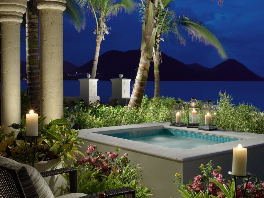 A poolside area with a view of the water and mountains, surrounded by palm trees and plants.