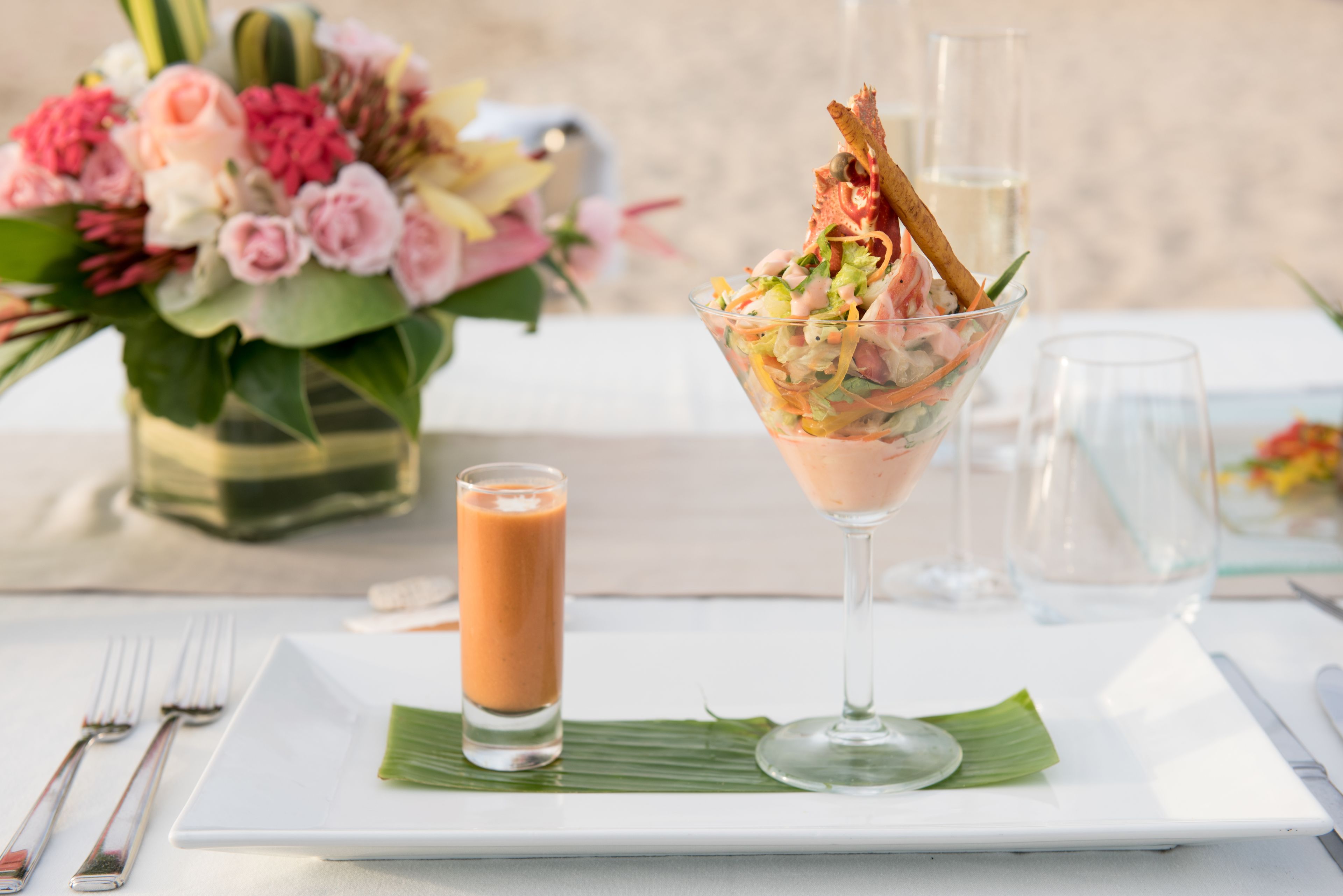 A table set with a glass of a pinkish drink, a clear glass with a pink drink, a plate with a green leaf, and a floral arrangement.